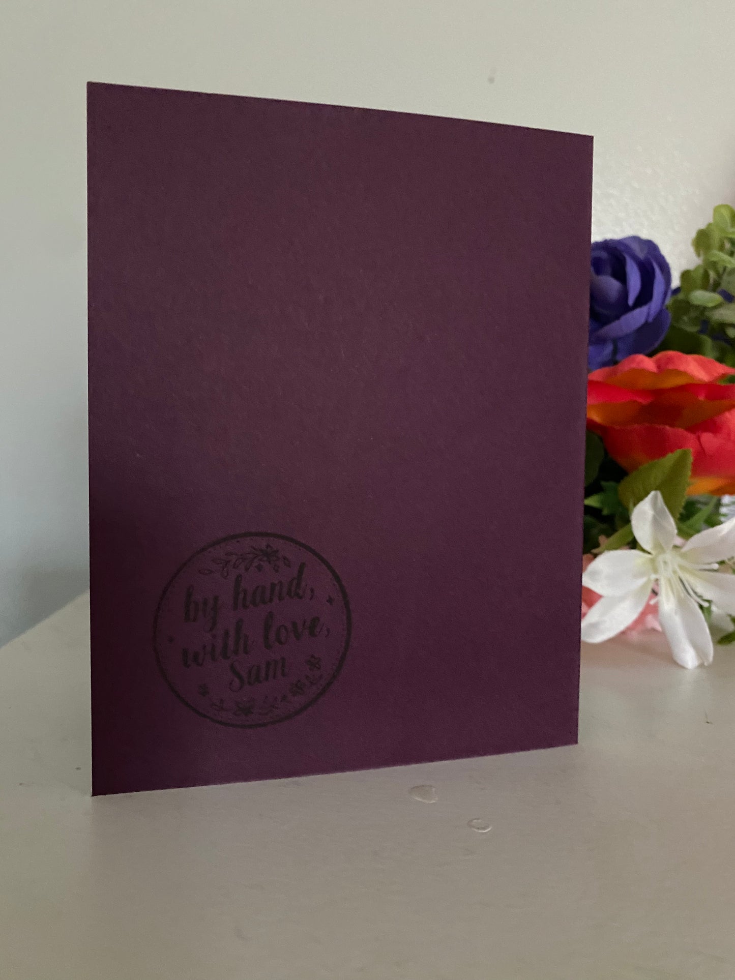 Purple card with 'by hand, with love' brand stamp, placed on a mauve or purple card.