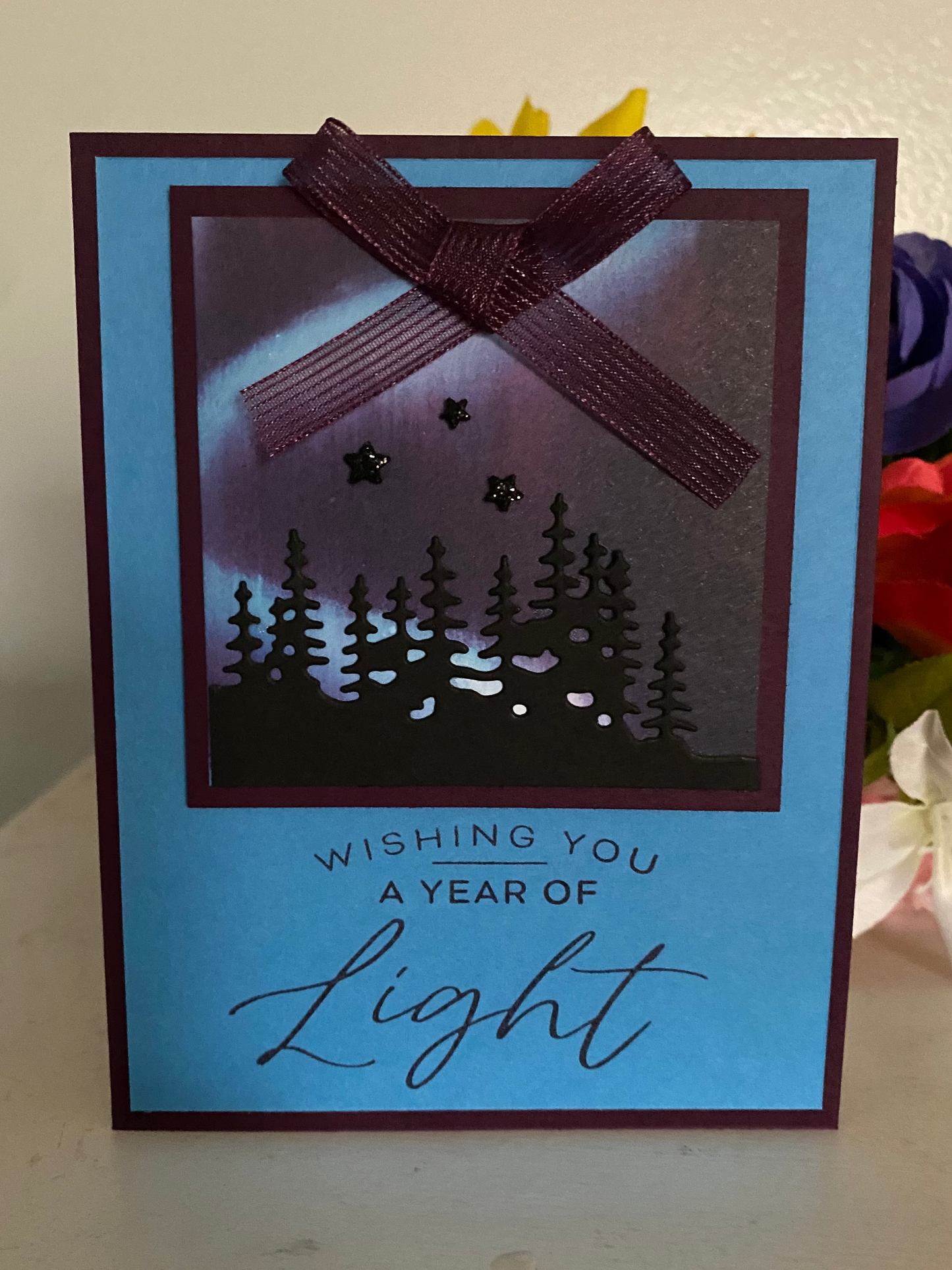 Greeting card with northern lights design and 'Wishing you a year of light' text, placed on a surface with northern lights in the background.
