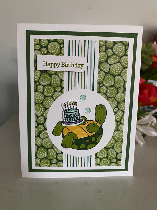 Birthday card with turtle holding a cake and 'Happy Birthday' text on a green patterned background.